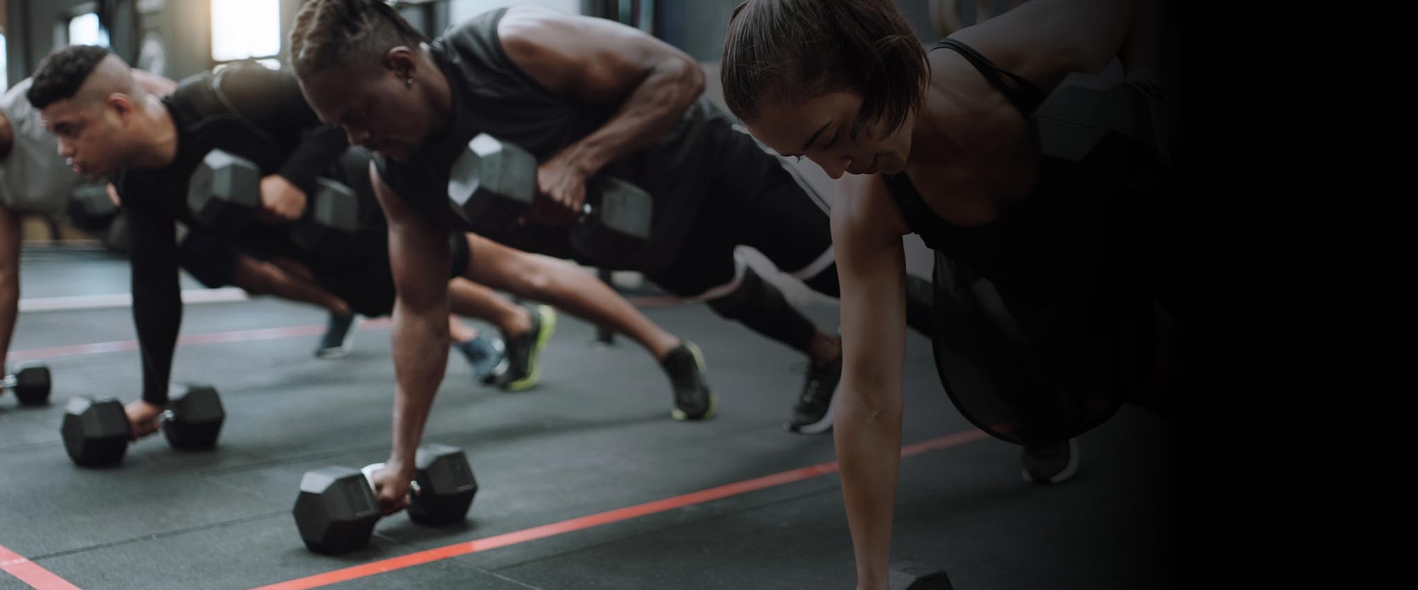 Header line up of people in work out class doing planks with weights.