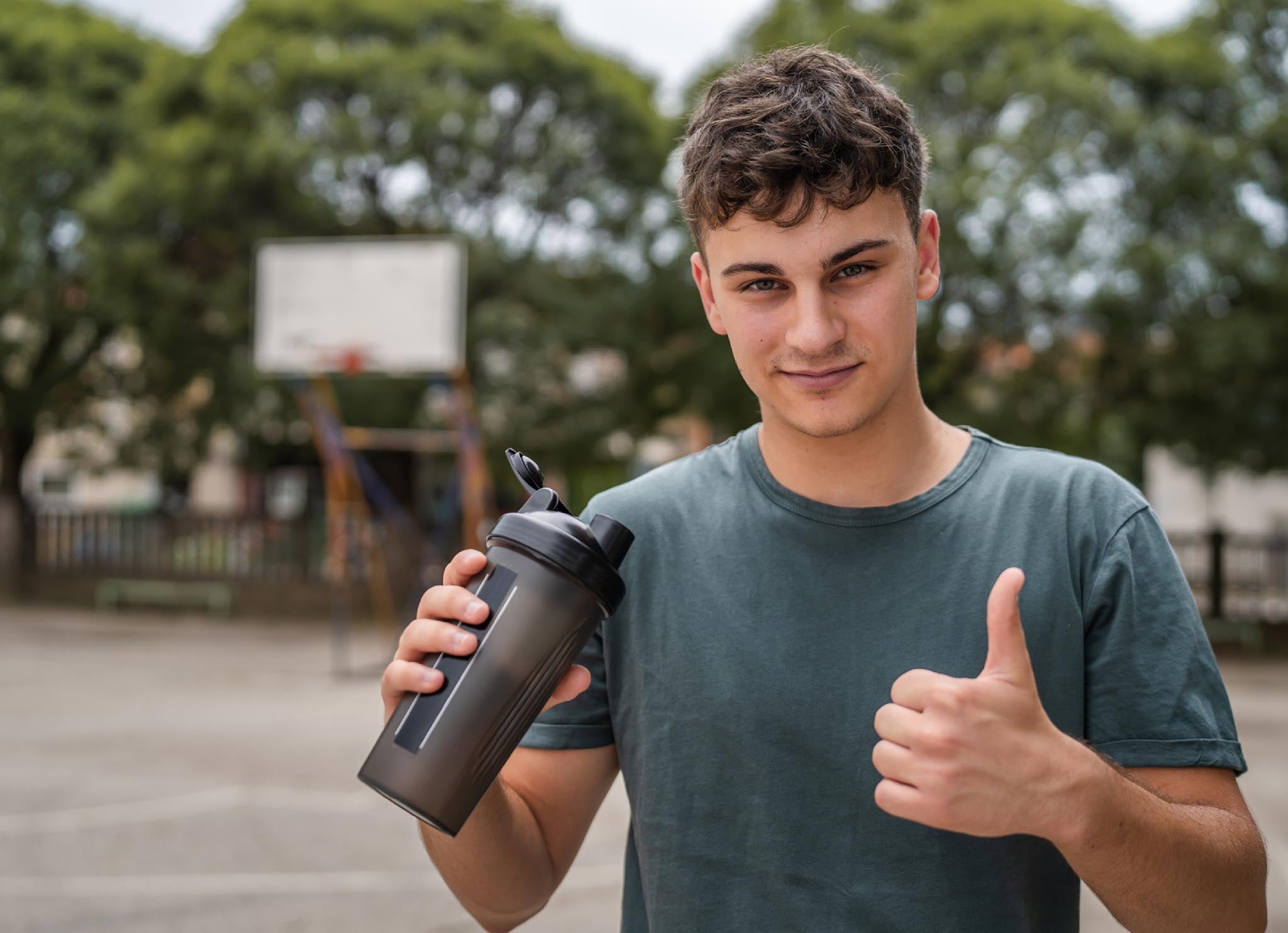 Young man in workout shirt with shaker bottle on outdoor basketball court giving thumbs up.