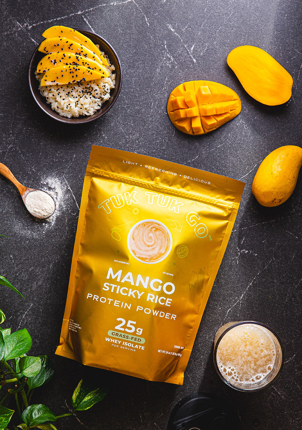 Mango sticky rice protein powder pouch top down shot with cut mango, mango and rice in bowl, spoon of powder, and glass of mixed protein.